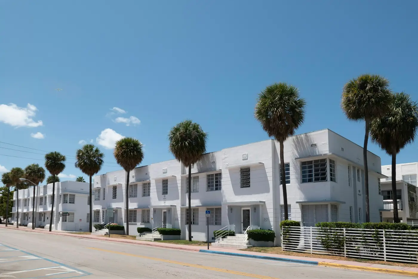 Row of white two-story Miami Beach apartment buildings with flat roofs and horizontal window bands, lined with sabal palms along the street