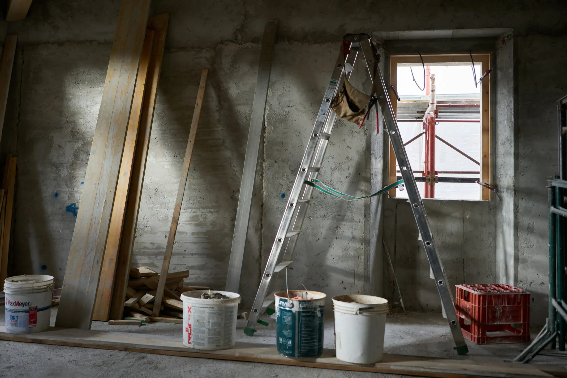Gutted concrete-block room mid-renovation, with an aluminum ladder beneath a window, paint buckets on the subfloor, and lumber leaning against the bare wall