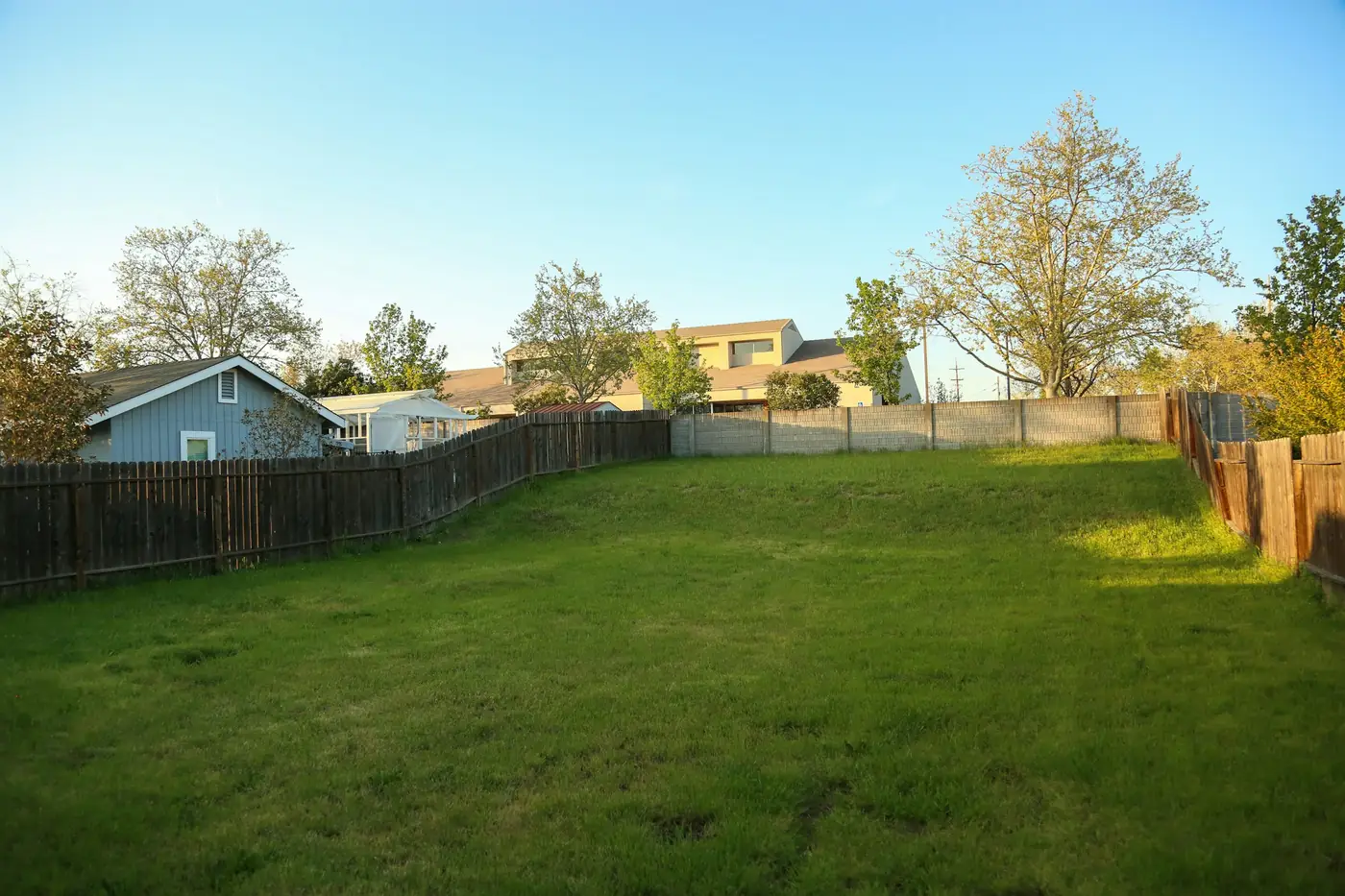 Empty grassy infill lot enclosed by wood privacy fences, with neighboring houses and trees along the back property line